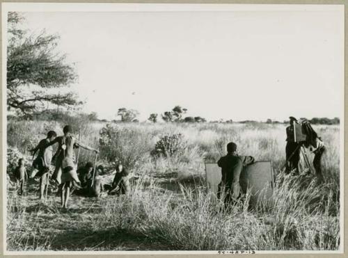 Oukwane, !Gai, /Gaiamakwe, !Ou/ke, and !Oukabe, working at a skin; /Twikwe, Tsekue, DaSi!Na, and N!whakwe dancing; expedition members filming the scene  (print is a cropped image)