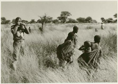 /Twikwe, DaSi!Na, and Tsekue carrying N!whakwe, walking in grass; William Donnelan taking a photo (print is a cropped image)