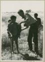 Tsekue, with N!whakwe on her back, standing beside John Marshall, DaSi!Na in the background (print is a cropped image)