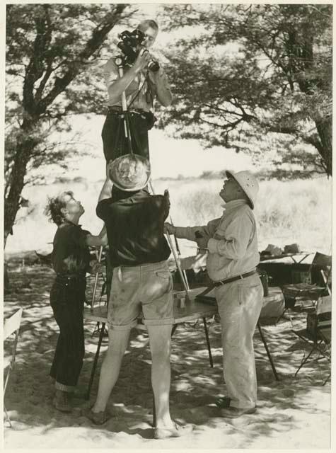 John Marshall standing on a table holding, a film camera; Lorna Marshall, Laurence Marshall, and an expedition member holding camera tripod steady (print is a cropped image)