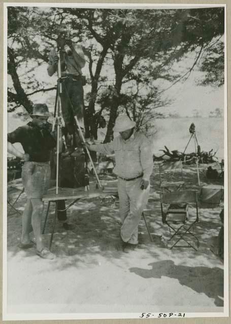 John Marshall standing on a table with a camera on a tripod, Laurence Marshall and an expedition member holding the tripod (print is a cropped image)