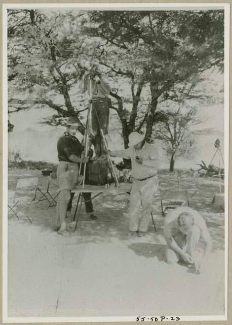 John Marshall standing on a table with a camera on a tripod, Laurence Marshall and an expedition member holding the tripod; another expedition member kneeling (print is a cropped image)