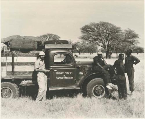 Expedition truck with sign painted on side reading "Peabody Museum of Harvard University," with Robert Story, Kernel Ledimo, /Gishay, and Simon Molamo gathered around

