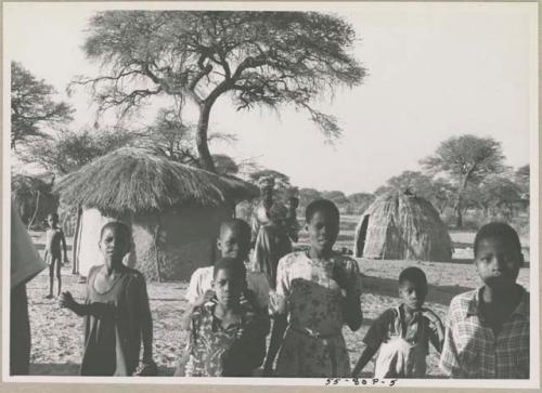 Group of women and children standing, with huts in background
