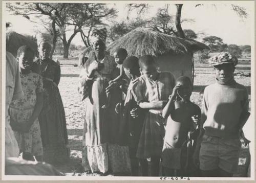 Group of people standing, with huts in background
