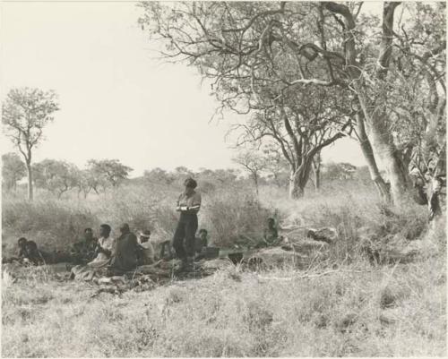 Elizabeth Marshall Thomas standing and taking notes with unidentified Ju/'hoansi seated around her









