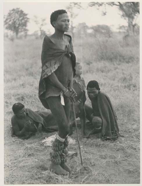 Young man standing, showing rattles and stick, with "/Qui Navel" and "Little ≠Gao," and somebody else seen in the background
