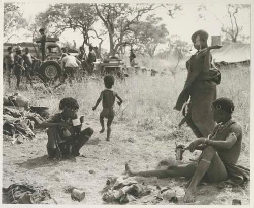 !U and N!ai, seated, an unidentified, visiting girl, standing, and !Ungka Norna walking away; unidentified people in the background around the expedition truck


























