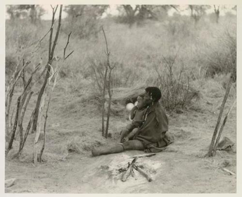 Woman seated by her fire, holding a child, seen from a distance























