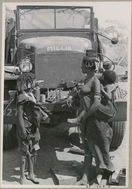 N!ai and a woman carrying a baby on her back, standing in front of one of the expedition's GMC trucks
























