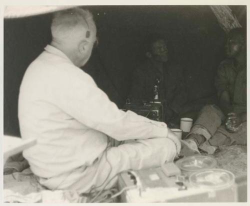 Laurence Marshall with earphones, conversing with ≠Toma and another man inside of a tent






















