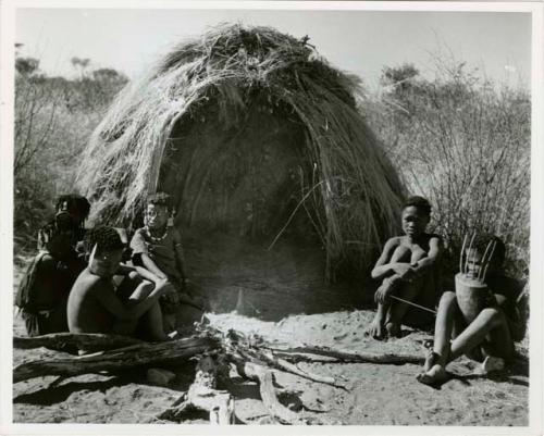 "N!ai and /Gunda": Little N!ai" and /Gunda (son of Khuan//a and her former husband) sitting by their skerm with their wedding party, including Tsamgao (son of ≠Toma and !U), ≠Gisa (daughter of ≠Gao and Khwo//o-/Gasa), Xama (daughter of "Gao Helmet" and //Kushay), ≠Gao (son of Gau and Be), and a boy playing the //guashi (print is a cropped image)