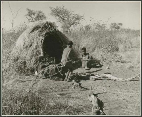 "N!ai and /Gunda": "Little N!ai" and /Gunda (son of Khuan//a and her former husband) sitting by their skerm with their wedding party, including Tsamgao (son of ≠Toma and !U), ≠Gisa (daughter of ≠Gao and Khwo//o-/Gasa), Xama (daughter of "Gao Helmet" and //Kushay), ≠Gao (son of Gau and Be), and a boy playing the //guashi (print is a cropped image)