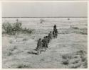 "Band moving": Band of people walking in a line through grass at the edge of a dry pan, view from behind (print is a cropped image)