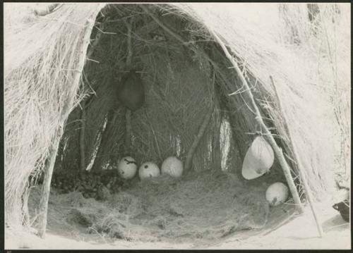 "Waterhole / Gwia": Interior of a skerm, with mangetti nuts, two water bags made from animal stomachs, ostrich egg shells, and grass used for bedding