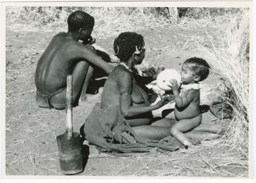 "Waterhole / Gwia": !Ungka (≠Toma's sister) with her daughter, Sa≠gai, sitting on her knees drinking from an ostrich eggshell; Tsamgao (Bau's husband) sitting beside her (print is a cropped image)