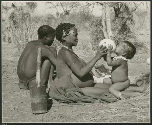 "Waterhole / Gwia": !Ungka (≠Toma's sister) with her daughter, Sa≠gai, sitting on her knees drinking from an ostrich eggshell; Tsamgao (Bau's husband) sitting beside her (print is a cropped image)