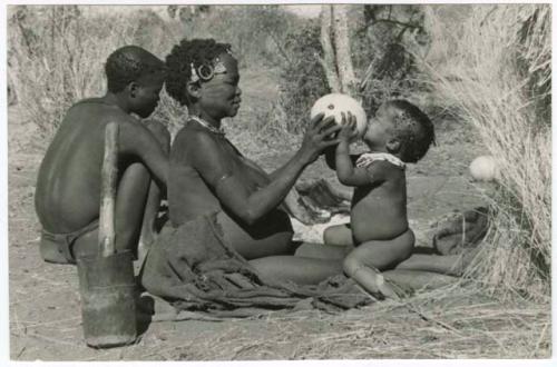 "Waterhole / Gwia": !Ungka (≠Toma's sister) with her daughter, Sa≠gai, sitting on her knees drinking from an ostrich eggshell; Tsamgao (Bau's husband) sitting beside her (print is a cropped image)