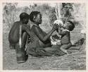 "Waterhole / Gwia": !Ungka (≠Toma's sister) with her daughter, Sa≠gai, sitting on her knees drinking from an ostrich eggshell; Tsamgao (Bau's husband) sitting beside her (print is a cropped image)