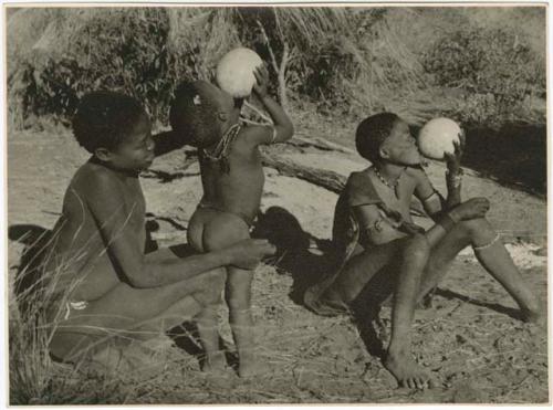 "Waterhole / Gwia": /Gunda (N!ai's husband) steadies a child while the child drinks from an ostrich eggshell, and a woman sitting next to them also drinking from a shell (print is a cropped image)
