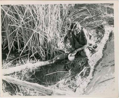 "Waterhole / Gwia": Woman filling an ostrich eggshell with water at a waterhole (print is a cropped image)