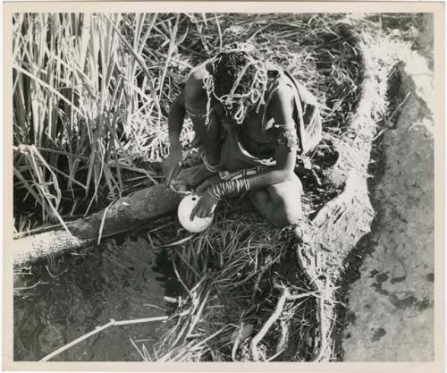 "Waterhole / Gwia": Woman filling an ostrich eggshell with water at a waterhole (print is a cropped image)