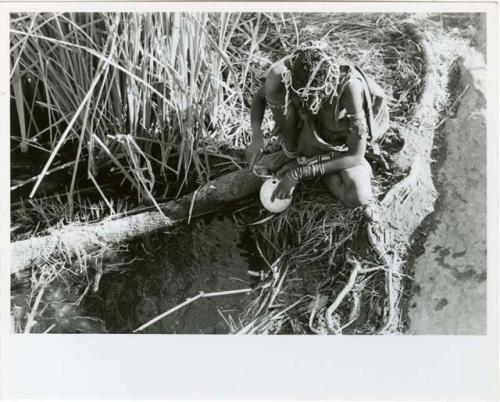 "Waterhole / Gwia": Woman filling an ostrich eggshell with water at a waterhole (print is a cropped image)