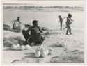 "Waterhole / Gwia": Khuan//a (Gau's wife) sitting with ostrich egg shells and children playing in the water behind her (print is a cropped image)