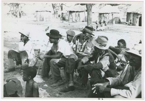 [No folder title]: Group of men sitting and watching a dance; huts in the background (print is a cropped image)