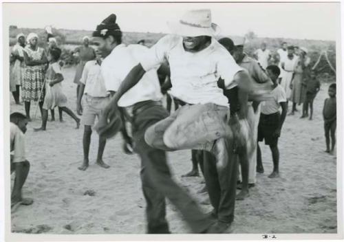 [No folder title]: Boy jumping over another boy's leg, playing a game in a school yard; people standing and watching in the background (print is a cropped image)