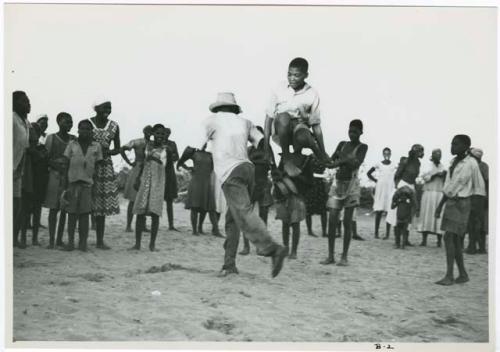 [No folder title]: Boy jumping over another boy's leg, playing a game in a school yard; people standing and watching in the background (print is a cropped image)