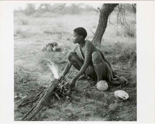 "'E', Chapter 9, 51": Woman squatting by fire, a melon beside her, settled for the night (print is a cropped image)