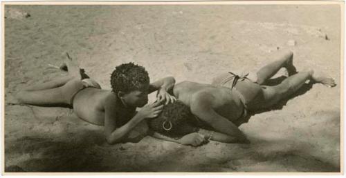 "Grooming & haircuts": Boy and man lying on the ground, the boy looking for lice in the man's hair (print is a cropped image)