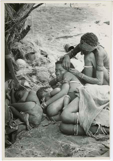 "Grooming & haircuts": Elderly woman adjusting the earring of a younger woman who is lying with her baby; another child asleep beside them (print is a cropped image)