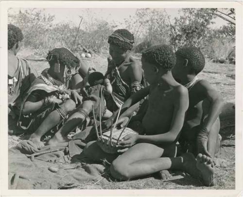 "Reel 224, 18-36, Ex III / b/w prints made from color slides, huddle": Group of people sitting, including "Little N!ai" (left), //Khuga (middle), and /Gishay (Gau's son) playing the //guashi (print is a cropped image)