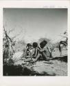 "Reel 224, 18-36, Ex III / b/w prints made from color slides, huddle": Group of people sitting, including "Little N!ai" (left), //Khuga (middle), and /Gishay (Gau's son) playing the //guashi (print is a cropped image)