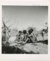 "Reel 224, 18-36, Ex III / b/w prints made from color slides, huddle": Group of people sitting, including "Little N!ai" (left), //Khuga (middle), and /Gishay (Gau's son) playing the //guashi (print is a cropped image)