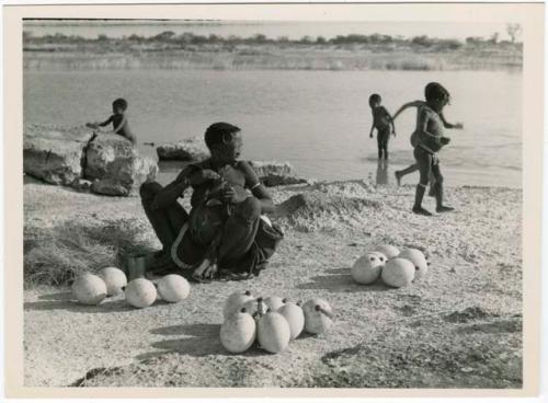 "Pan": Khuan//a (Gau's wife) sitting with ostrich egg shells and children playing in the water behind her (print is a cropped image)