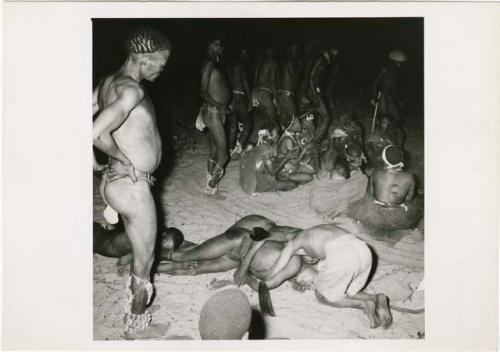 [No folder title]: Three medicine men lying on the ground in trance, with //Gunda kneeling over one of them, men dancing around a circle of women sitting and clapping in the background (print is a cropped image)