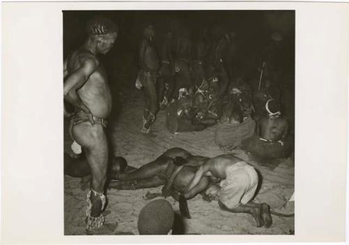 [No folder title]: Three medicine men lying on the ground in trance, with //Gunda kneeling over one of them, men dancing around a circle of women sitting and clapping in the background (print is a cropped image)