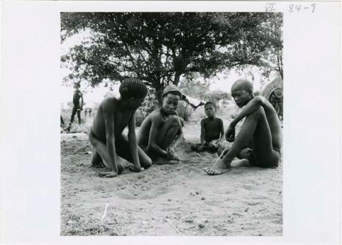 [No folder title]: Four boys sitting, playing /Ui (the counting game), with the boy on the right holding a slender white stick (print is a cropped image)