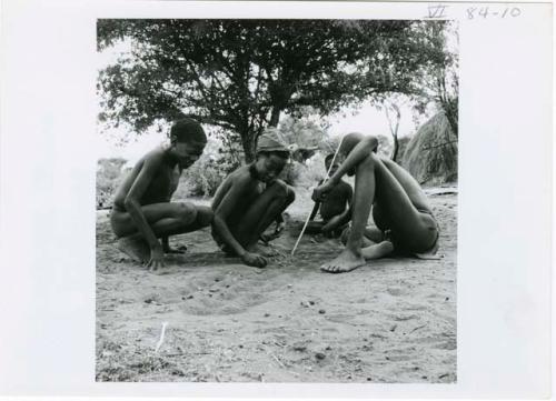 [No folder title]: Four boys sitting, playing /Ui (the counting game), with the boy on the right pushing pebbles with a stick (print is a cropped image)