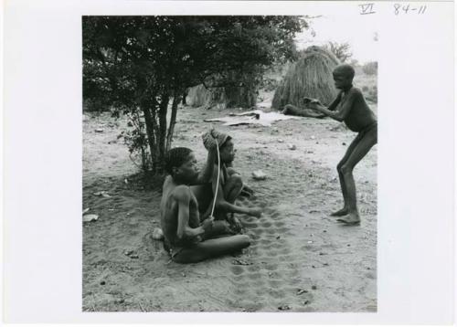 [No folder title]: Three boys sitting, playing /Ui (the counting game), with the boy on the right gesturing and the boy on the left holding a stick; skerms and animal skins on the ground next to them in the background (print is a cropped image)