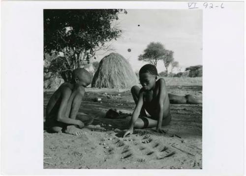 [No folder title]: Two boys playing /Ui (the counting game) in the sand, with a skerm in the background (print is a cropped image)