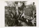 [No folder title]: Two medicine men sitting back to back on the ground at a daytime dance, with two men standing and looking at them (print is a cropped image)