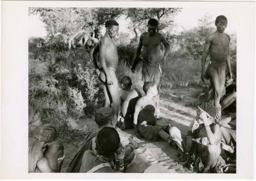 [No folder title]: Two medicine men sitting back to back on the ground at a daytime dance, with two men standing and looking at them (print is a cropped image)