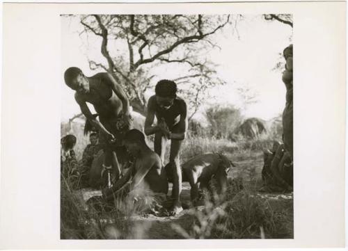 [No folder title]: /Gunda sitting on the ground in trance at a daytime dance, with a man kneeling in back of him, leaning his head on his back, /Ti!kay and another man standing near them (print is a cropped image)