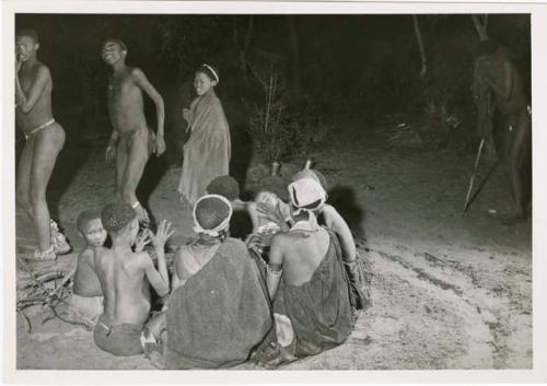 [No folder title]: Dancers dance by a group of women sitting and clapping in a night dance (print is a cropped image)