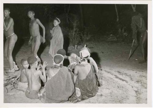 [No folder title]: Dancers dance by a group of women sitting and clapping in a night dance (print is a cropped image)