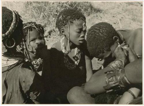 "Ornaments": Group of women and girls sitting, and one woman fixing another woman's earring (print is a cropped image)